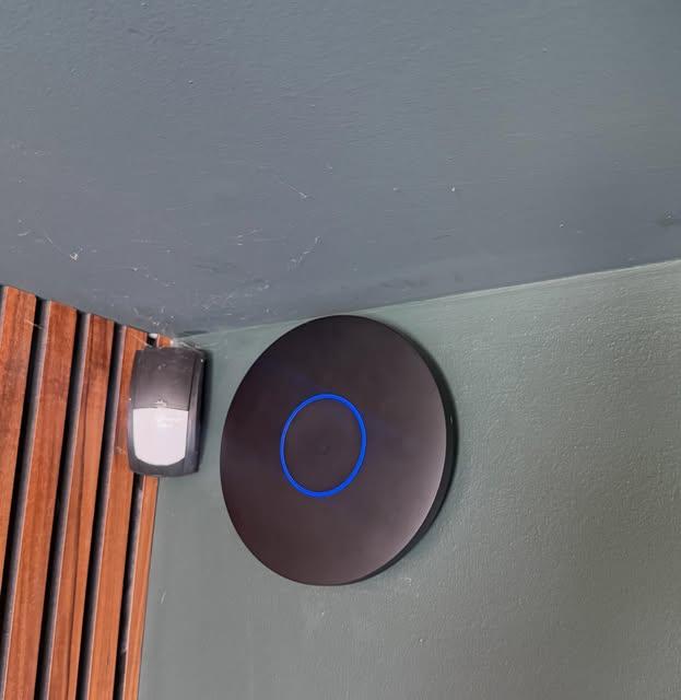 Black Ubiquiti UniFi WiFi access point with blue status ring mounted on a green interior wall